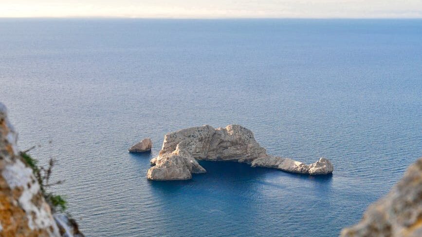 A serene aerial view of an uninhabited rock island in the Mediterranean Sea, surrounded by clear blue waters.